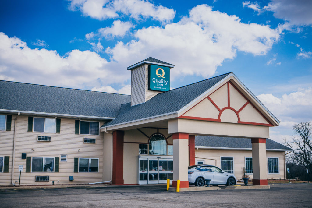 Photo of the entry to the Quality Inn hotel in Edgerton. The entry has a covered pull-up area showing a parked car. The green Quality Inn sign is shown high in the blue and cloudy sky